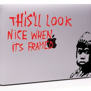 Puede incluir: Un portátil plateado con una pegatina roja y negra que dice "This'll look nice when it's framed" y una imagen en blanco y negro de una persona con un sombrero.