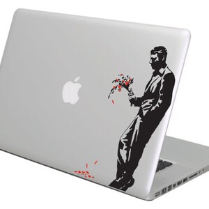 Puede incluir: Un portátil plateado con una pegatina negra y roja de un hombre sosteniendo flores. El hombre está silueteado sobre un fondo blanco. Las flores son rojas y los pétalos caen al suelo.