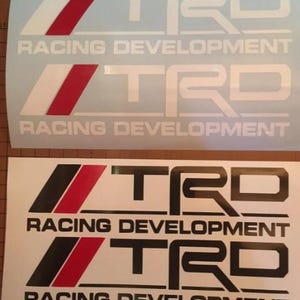 Könnte beinhalten: Zwei Sätze von TRD Racing Development Aufklebern. Der obere Satz hat weißen Text und einen weißen und roten Streifen. Der untere Satz hat schwarzen Text und einen schwarzen und roten Streifen. Die Aufkleber sind auf weißem Hintergrund.