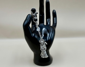 May include: A silver-toned metal keychain featuring a detailed charm of a hooded skeletal figure holding a scythe, suspended from a short link chain and a lobster claw clasp. The charm includes a circular halo detail behind the skull.