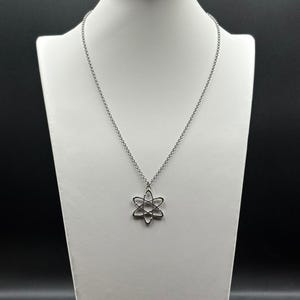 K&ouml;nnte beinhalten: Eine silberfarbene Halskette mit einem Anh&auml;nger in Form eines Atoms. Der Anh&auml;nger hat ein sechszackiges Stern-Design mit einem kreisf&ouml;rmigen Zentrum. Die Halskette wird auf einer wei&szlig;en Schaufensterpuppe pr&auml;sentiert.