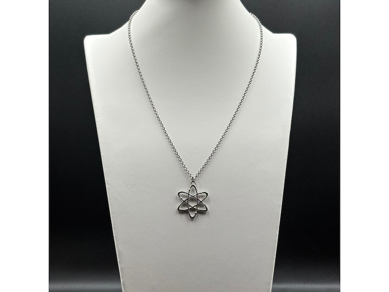 K&ouml;nnte beinhalten: Eine silberfarbene Halskette mit einem Anh&auml;nger in Form eines Atoms. Der Anh&auml;nger hat ein sechszackiges Stern-Design mit einem kreisf&ouml;rmigen Zentrum. Die Halskette wird auf einer wei&szlig;en Schaufensterpuppe pr&auml;sentiert.