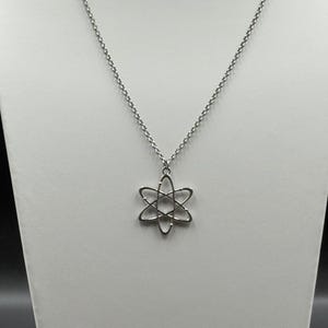 K&ouml;nnte beinhalten: Eine silberfarbene Halskette mit einem Anh&auml;nger in Form eines Atoms. Der Anh&auml;nger hat ein sechszackiges Stern-Design mit geschwungenen Linien. Die Halskette hat eine zarte, perlenbesetzte Kette. Der Schmuck wird auf einer wei&szlig;en Oberfl&auml;che pr&auml;sentiert.