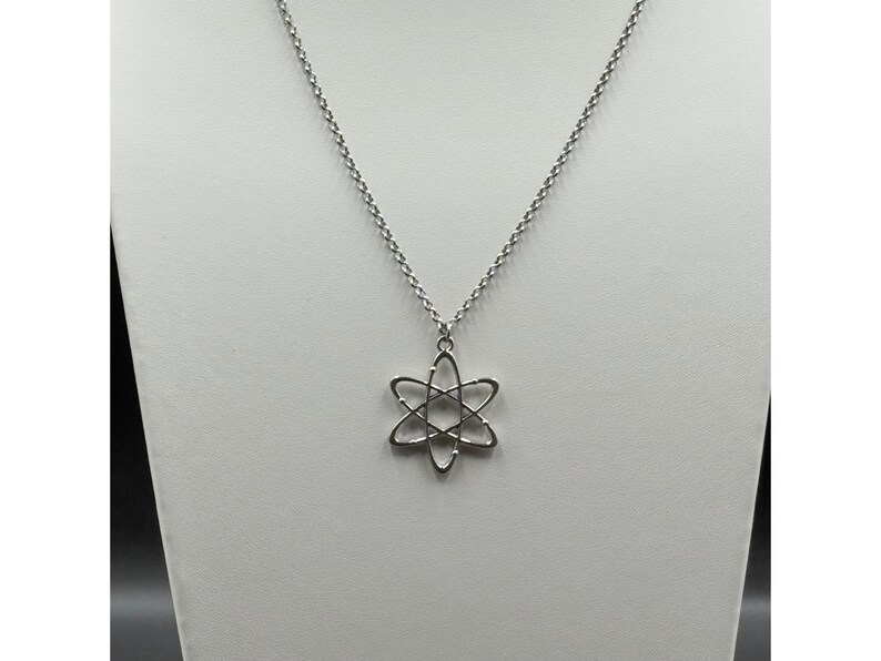 K&ouml;nnte beinhalten: Eine silberfarbene Halskette mit einem Anh&auml;nger in Form eines Atoms. Der Anh&auml;nger hat ein sechszackiges Stern-Design mit geschwungenen Linien. Die Halskette hat eine zarte, perlenbesetzte Kette. Der Schmuck wird auf einer wei&szlig;en Oberfl&auml;che pr&auml;sentiert.