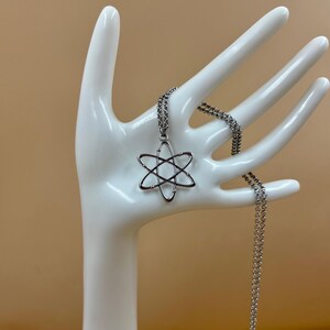 K&ouml;nnte beinhalten: Eine silberfarbene Halskette mit einem Atom-Anh&auml;nger. Der Anh&auml;nger hat ein sechszackiges Sterndesign, und die Kette ist im filigranen Glieder-Stil gehalten. Die Halskette wird auf einem wei&szlig;en, handf&ouml;rmigen Schmuckhalter pr&auml;sentiert.