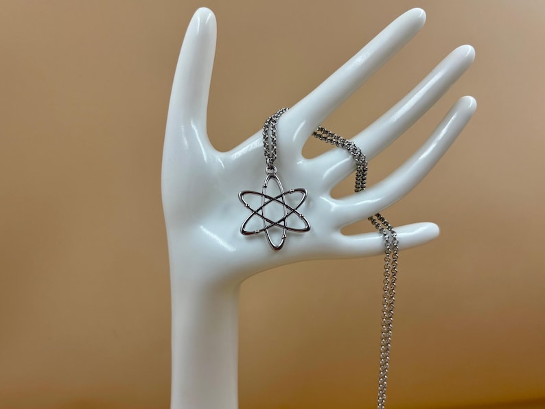 K&ouml;nnte beinhalten: Eine silberfarbene Halskette mit einem Atom-Anh&auml;nger. Der Anh&auml;nger hat ein sechszackiges Sterndesign, und die Kette ist im filigranen Glieder-Stil gehalten. Die Halskette wird auf einem wei&szlig;en, handf&ouml;rmigen Schmuckhalter pr&auml;sentiert.