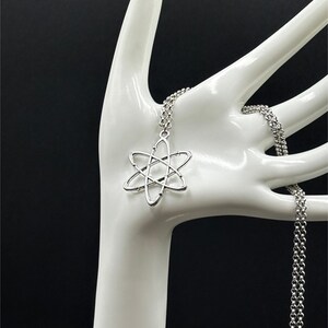 K&ouml;nnte beinhalten: Halskette mit einem silberfarbenen Anh&auml;nger in Form eines Atoms. Der Anh&auml;nger h&auml;ngt an einer feinen Kette. Die Halskette wird auf einer wei&szlig;en, handf&ouml;rmigen Halterung vor schwarzem Hintergrund pr&auml;sentiert.