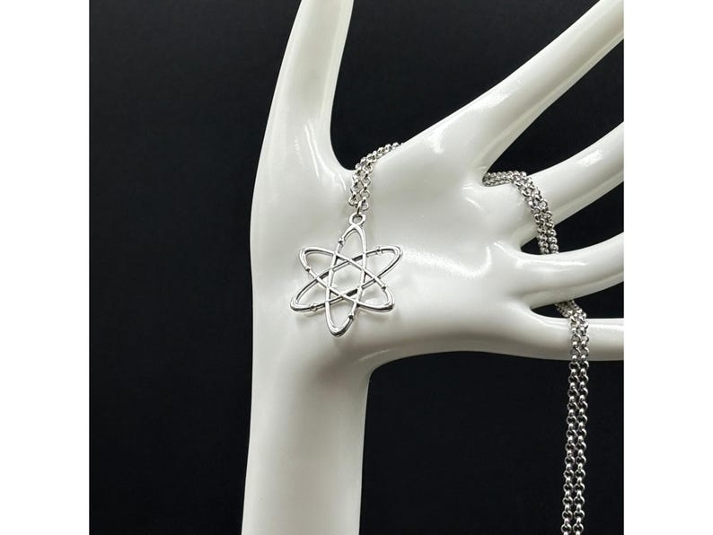 K&ouml;nnte beinhalten: Halskette mit einem silberfarbenen Anh&auml;nger in Form eines Atoms. Der Anh&auml;nger h&auml;ngt an einer feinen Kette. Die Halskette wird auf einer wei&szlig;en, handf&ouml;rmigen Halterung vor schwarzem Hintergrund pr&auml;sentiert.