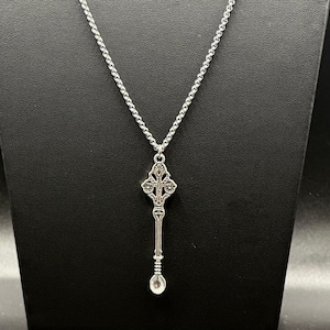 May include: A silver-toned necklace featuring a long, slender pendant shaped like a decorative spoon or wand. The pendant has an intricate, symmetrical filigree design at the top and a small, rounded bowl at the bottom, suspended from a silver chain.