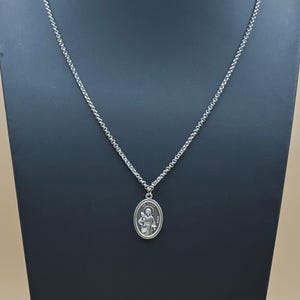 May include: A silver-toned necklace with a detailed oval pendant featuring a religious figure. The pendant hangs from a delicate chain. The necklace is displayed on a black stand against a neutral background.