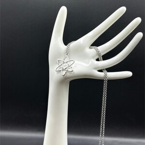 K&ouml;nnte beinhalten: Eine silberfarbene Halskette mit einem Atom-Anh&auml;nger. Der Anh&auml;nger ist eine stilisierte Darstellung der Struktur eines Atoms. Die Halskette wird auf einem wei&szlig;en, handf&ouml;rmigen Schmuckhalter vor schwarzem Hintergrund pr&auml;sentiert.