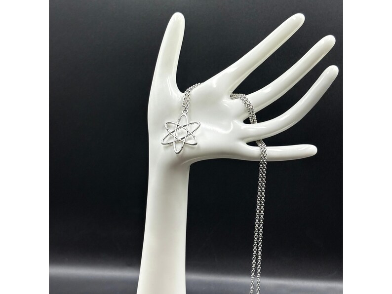 K&ouml;nnte beinhalten: Eine silberfarbene Halskette mit einem Atom-Anh&auml;nger. Der Anh&auml;nger ist eine stilisierte Darstellung der Struktur eines Atoms. Die Halskette wird auf einem wei&szlig;en, handf&ouml;rmigen Schmuckhalter vor schwarzem Hintergrund pr&auml;sentiert.