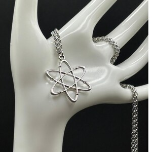 K&ouml;nnte beinhalten: Silberfarbene Halskette mit einem Anh&auml;nger in Atomform. Der Anh&auml;nger hat einen Mittelpunkt mit sechs umlaufenden Linien. Die Kette ist ebenfalls silberfarben und hat ein Glieddesign. Die Halskette wird auf einer wei&szlig;en Hand pr&auml;sentiert.