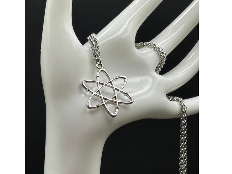 K&ouml;nnte beinhalten: Silberfarbene Halskette mit einem Anh&auml;nger in Atomform. Der Anh&auml;nger hat einen Mittelpunkt mit sechs umlaufenden Linien. Die Kette ist ebenfalls silberfarben und hat ein Glieddesign. Die Halskette wird auf einer wei&szlig;en Hand pr&auml;sentiert.
