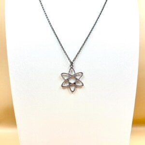 K&ouml;nnte beinhalten: Eine silberfarbene Halskette mit einem sechszackigen Sternanh&auml;nger. Der Anh&auml;nger hat ein offenes, geometrisches Design und h&auml;ngt an einer feinen Kette. Die Halskette wird auf einer wei&szlig;en Schaufensterpuppe pr&auml;sentiert.