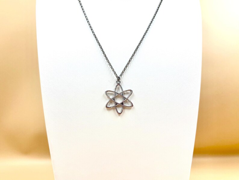 K&ouml;nnte beinhalten: Eine silberfarbene Halskette mit einem sechszackigen Sternanh&auml;nger. Der Anh&auml;nger hat ein offenes, geometrisches Design und h&auml;ngt an einer feinen Kette. Die Halskette wird auf einer wei&szlig;en Schaufensterpuppe pr&auml;sentiert.