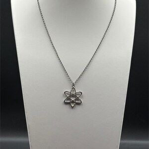 K&ouml;nnte beinhalten: Eine silberfarbene Halskette mit einem Anh&auml;nger in Form eines Atoms. Der Anh&auml;nger hat ein sechszackiges Sterndesign mit einem kreisf&ouml;rmigen Zentrum. Die Halskette wird auf einer wei&szlig;en B&uuml;ste vor dunklem Hintergrund pr&auml;sentiert.