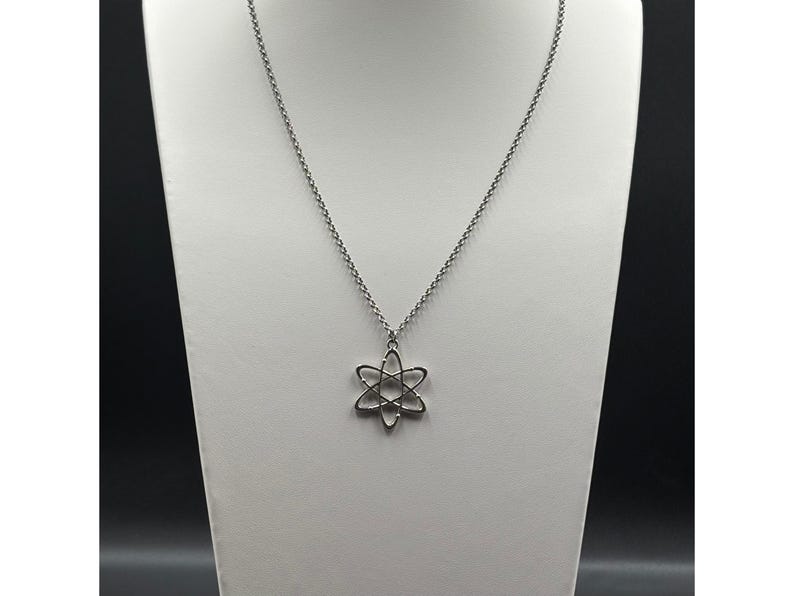 K&ouml;nnte beinhalten: Eine silberfarbene Halskette mit einem Anh&auml;nger in Form eines Atoms. Der Anh&auml;nger hat ein sechszackiges Sterndesign mit einem kreisf&ouml;rmigen Zentrum. Die Halskette wird auf einer wei&szlig;en B&uuml;ste vor dunklem Hintergrund pr&auml;sentiert.