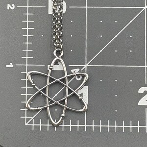 K&ouml;nnte beinhalten: Silberfarbene Atom-Anh&auml;nger-Halskette. Der Anh&auml;nger hat ein detailliertes Atomdesign mit umlaufenden Elektronen. Die Halskette h&auml;ngt an einer filigranen Kette. Der Hintergrund ist eine graue Oberfl&auml;che mit Messmarkierungen.