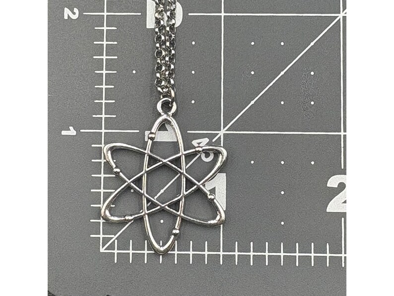 K&ouml;nnte beinhalten: Silberfarbene Atom-Anh&auml;nger-Halskette. Der Anh&auml;nger hat ein detailliertes Atomdesign mit umlaufenden Elektronen. Die Halskette h&auml;ngt an einer filigranen Kette. Der Hintergrund ist eine graue Oberfl&auml;che mit Messmarkierungen.