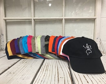 May include: A collection of baseball caps in various colors, including black, blue, pink, and green, are displayed on a white wooden surface. The black cap in the foreground features a white embroidered skeleton design. The caps are arranged in a row.
