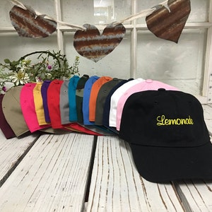 May include: A collection of 15 blank baseball caps in various colors, including pink, red, yellow, green, blue, and black. The black cap has the word "Lemonade" embroidered in yellow on the front.
