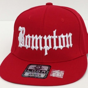 May include: Red baseball cap with a white embroidered "Compton" in old English lettering. The cap has a black and white label that reads "L.O.G.A. Leader of Generations" and "Size One Size Fits All."
