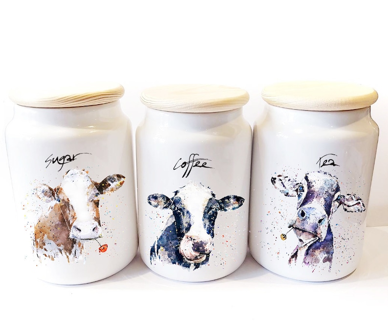 May include: Three white ceramic canisters with wooden lids, each featuring a watercolor illustration of a cow. The canisters are labeled "Sugar", "Coffee", and "Tea".