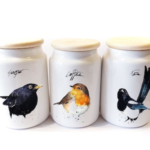 以下が含まれることがあります： 木製の蓋が付いた白いセラミック製の容器が3つ。それぞれの容器には異なる鳥の絵と、「砂糖」、「コーヒー」、「紅茶」の文字が書かれています。