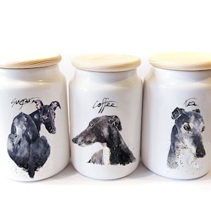 May include: Three white ceramic canisters with wooden lids. Each canister features a watercolor illustration of a greyhound dog and the words "Sugar", "Coffee", and "Tea" in black script. The canisters are designed for kitchen storage.