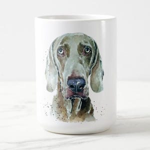 Peut inclure: Mug en céramique blanche avec une peinture à l'aquarelle d'un chien Weimaraner. Le chien a un pelage gris, des yeux bleus et un nez brun. La tasse est cylindrique et a une finition brillante. L'œuvre est détaillée et réaliste.