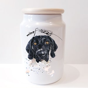 May include: White ceramic dog treat jar with a wooden lid. The jar features a black and white illustration of a dog's face with the words "woof treats" written in black cursive.
