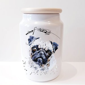 May include: White ceramic dog treat jar with a wooden lid. The jar features a watercolour illustration of a pug with the words "woof treats" written on the front.
