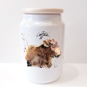 Pu&ograve; includere: Un barattolo di ceramica bianca per il caff&egrave; con un coperchio di legno. Il barattolo presenta un'illustrazione ad acquerello di un vitello marrone e la parola "caff&egrave;" in corsivo nero.