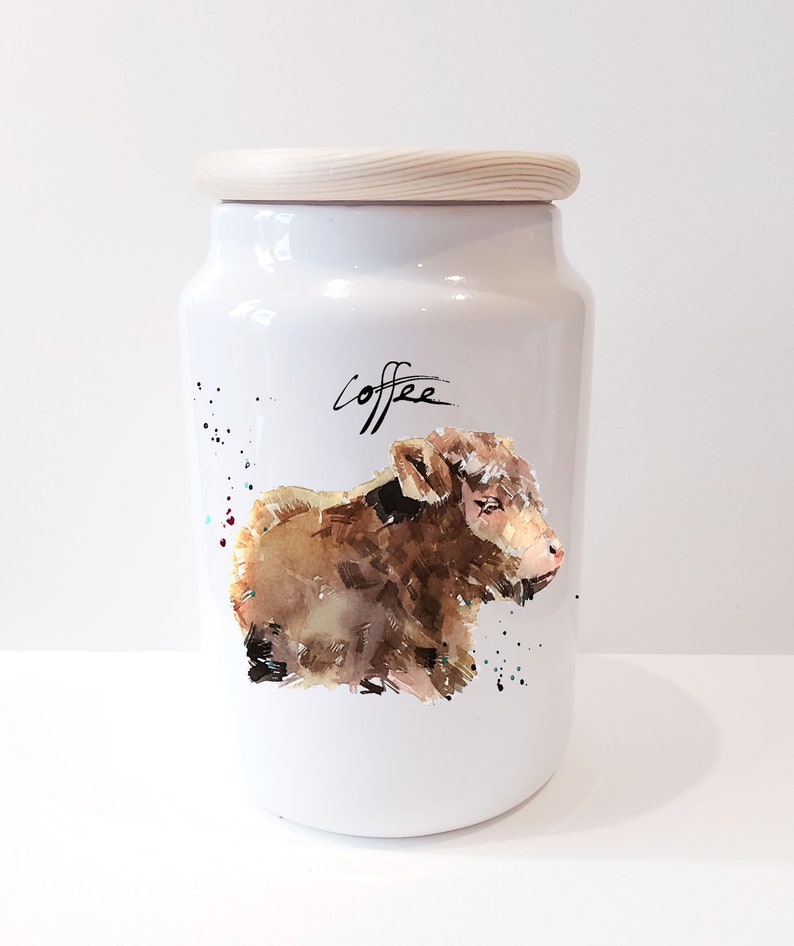 Pu&ograve; includere: Un barattolo di ceramica bianca per il caff&egrave; con un coperchio di legno. Il barattolo presenta un'illustrazione ad acquerello di un vitello marrone e la parola "caff&egrave;" in corsivo nero.