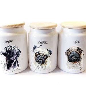 May include: Three white ceramic canisters with wooden lids, each featuring a different pug illustration. The canisters are labelled "Sugar", "Coffee", and "Tea".