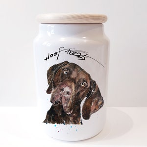 May include: White ceramic dog treat jar with a wooden lid. The jar features a brown and white illustration of a dog's head with the words "woof treats" written in black cursive.