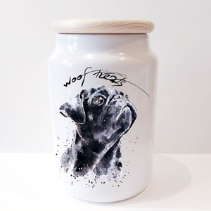 May include: White ceramic dog treat jar with a wooden lid. The jar features a black and white watercolour illustration of a pug with the text "woof treats" written on the front.
