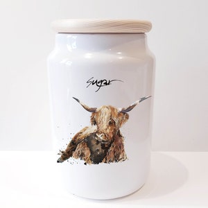 Pu&ograve; includere: Barattolo di zucchero in ceramica bianca con coperchio in legno. Il barattolo presenta un'illustrazione ad acquerello di una mucca Highland marrone con la faccia bianca. La parola "Zucchero" &egrave; scritta in corsivo nero sulla parte anteriore del barattolo.