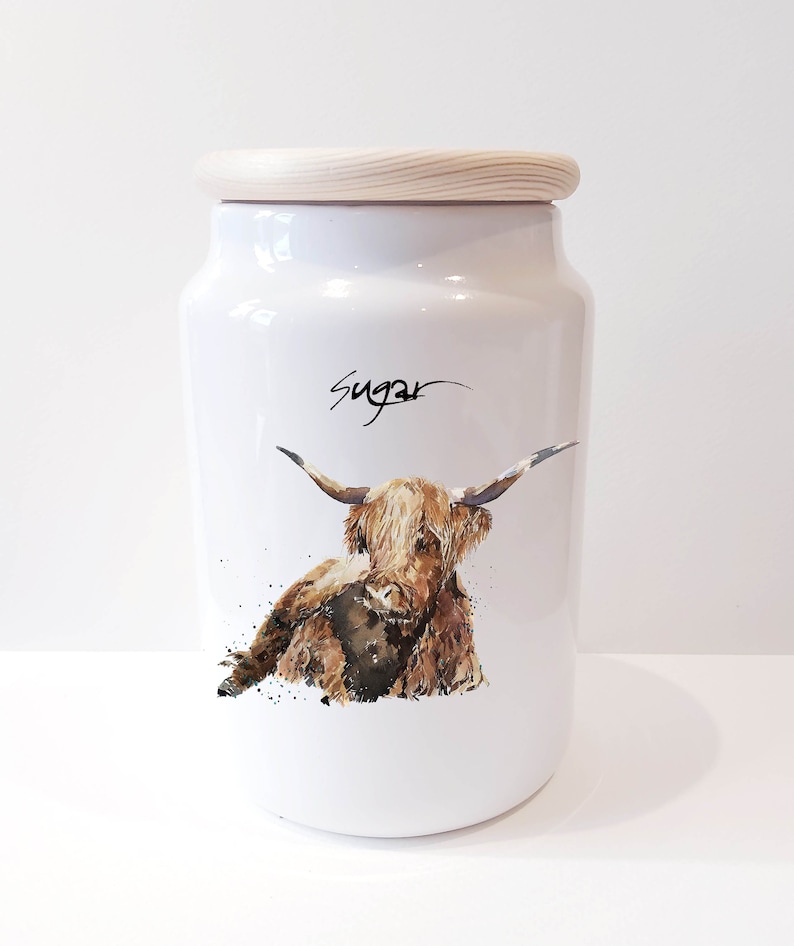 Pu&ograve; includere: Barattolo di zucchero in ceramica bianca con coperchio in legno. Il barattolo presenta un'illustrazione ad acquerello di una mucca Highland marrone con la faccia bianca. La parola "Zucchero" &egrave; scritta in corsivo nero sulla parte anteriore del barattolo.