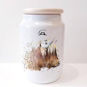 Pu&ograve; includere: Un barattolo di t&egrave; in ceramica bianca con un coperchio in legno. Il barattolo presenta un'illustrazione ad acquerello di una mucca delle Highlands con pelo marrone e viso bianco. La parola "T&egrave;" &egrave; scritta in corsivo nero sul davanti del barattolo.