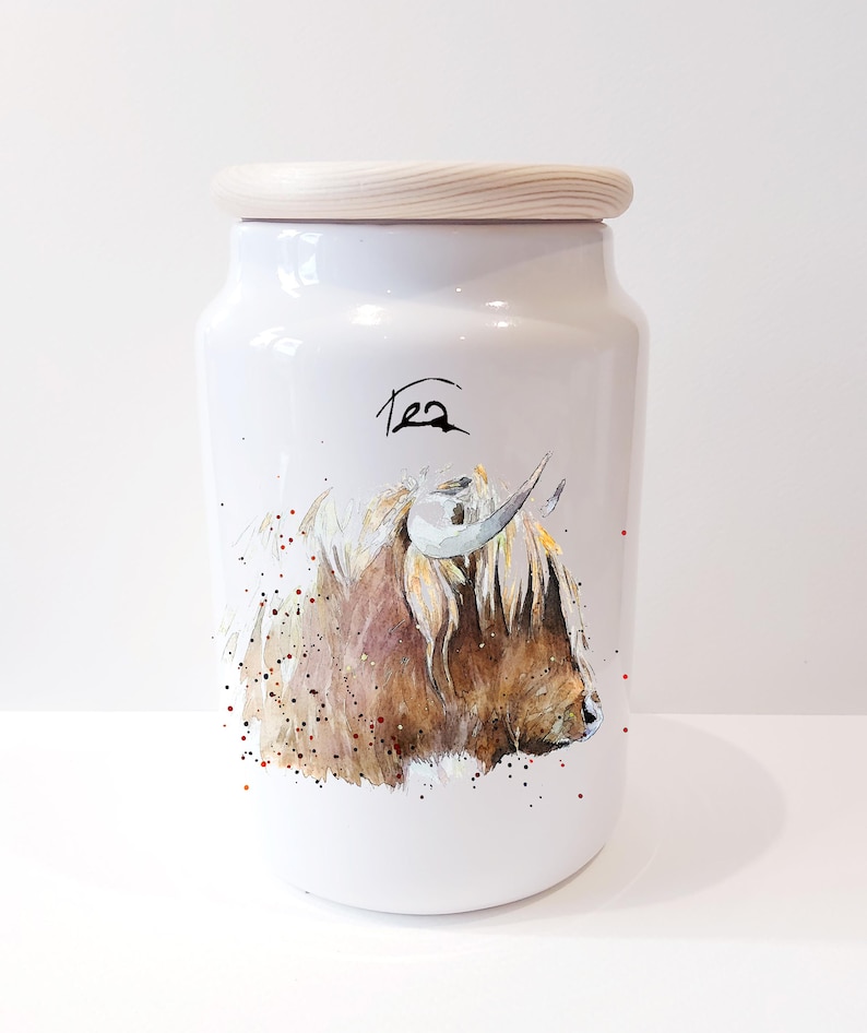Pu&ograve; includere: Un barattolo di t&egrave; in ceramica bianca con un coperchio in legno. Il barattolo presenta un'illustrazione ad acquerello di una mucca delle Highlands con pelo marrone e viso bianco. La parola "T&egrave;" &egrave; scritta in corsivo nero sul davanti del barattolo.