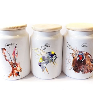 Può includere: Set di tre barattoli in ceramica bianca con coperchi in legno. I barattoli sono decorati con illustrazioni ad acquerello di una lepre, un bombo e un fagiano. I barattoli sono etichettati "Zucchero", "Caffè" e "Tè".