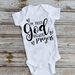 May include: White baby onesie with black text that reads "I'm proof God answers prayers" with hearts around the text.