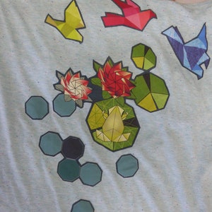 May include: A light blue t-shirt with a geometric design featuring a frog, flowers, and birds. The design is in shades of green, red, blue, and yellow.