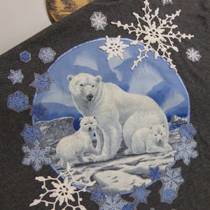 Pode incluir: Uma camiseta cinza com uma imagem azul e branca de ursos polares em uma paisagem nevada. A camiseta é decorada com flocos de neve de crochê branco e apliques em forma de floco de neve azul.