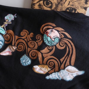 Op de afbeelding: Zwart T-shirt met geborduurde patches van een zeemeermin, schelpen en een golf met een boot. De patches zijn in tinten blauw, bruin en roze.