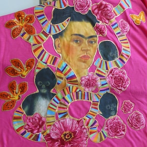 May include: A pink t-shirt with a colorful, striped ribbon design. The ribbon design surrounds a portrait of Frida Kahlo, two monkeys, and various flowers.