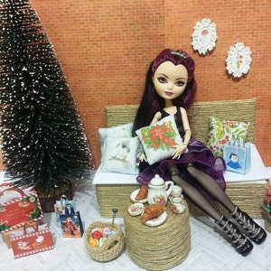 May include: A doll in a black dress and purple skirt sits on a woven couch with pillows. A small table holds a tea set and pastries. A Christmas tree and gift bags are nearby, creating a festive holiday scene.