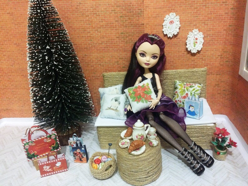 May include: A doll in a black dress and purple skirt sits on a woven couch with pillows. A small table holds a tea set and pastries. A Christmas tree and gift bags are nearby, creating a festive holiday scene.